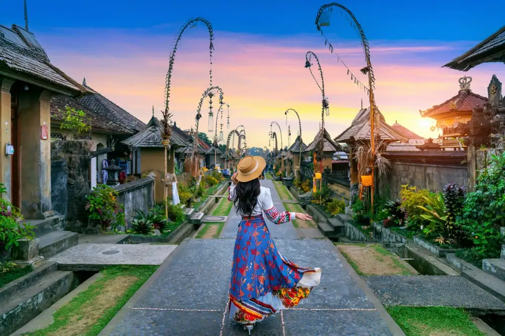 A woman walks down a traditional Balinese stone street under a sunset sky.
