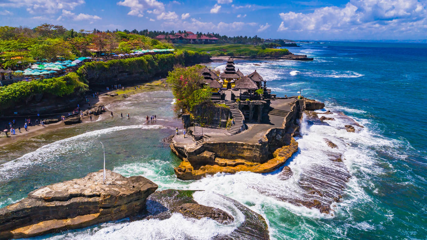 Tanah Lot, Bali - Aerial birdview of a historic seaside temple on a rocky outcrop surrounded by turquoise ocean waters and lush green coastline under a bright blue sky.