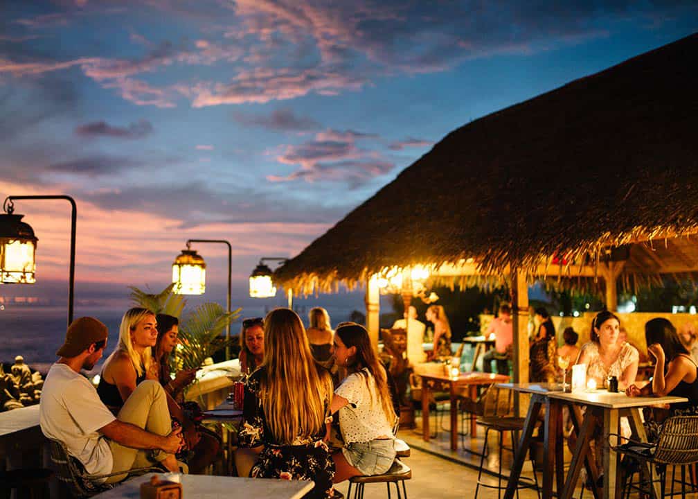 Beachfront outdoor lounge terrace at sunset with wooden armchairs, blue cushions, glowing lanterns, and scenic ocean views under shade trees.