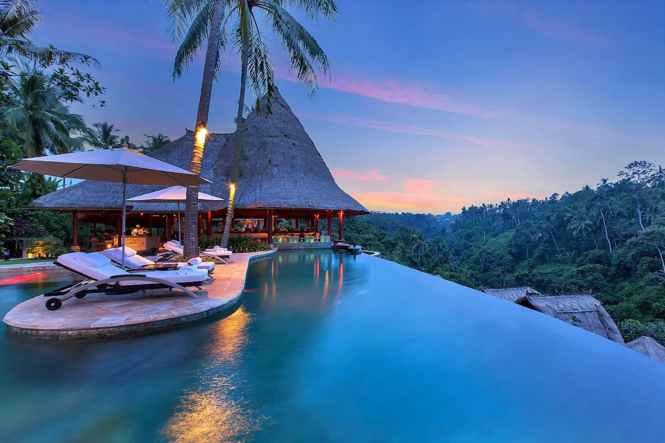 A luxury infinity pool overlooking a lush green tropical valley at sunset with lounge chairs and thatched-roof pavilions.