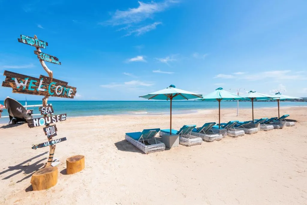 A scenic beach front area with light blue loungers, umbrellas, a rustic wooden welcome sign, and clear blue ocean waters.
