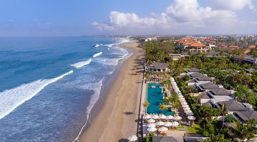 Aerial view of a luxury oceanfront resort featuring a long turquoise swimming pool, sandy beach, and rolling waves.