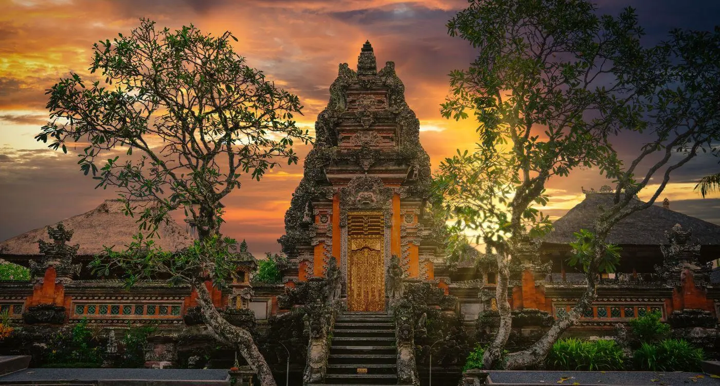 Traditional Balinese temple gate with intricate stone carvings and a golden door, framed by trees against a vibrant sunset sky.
