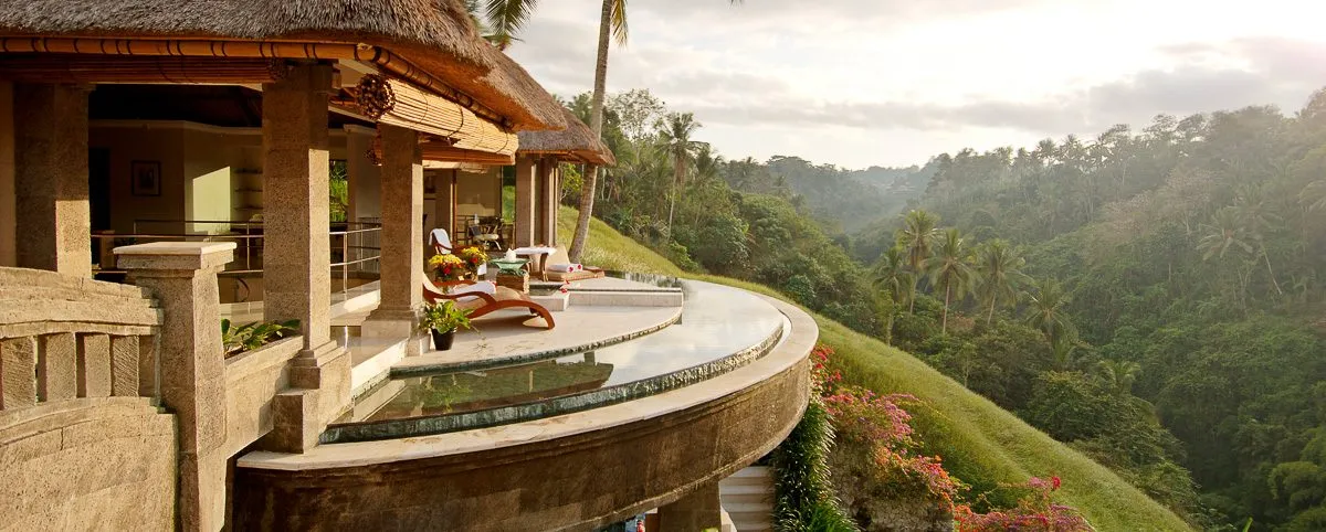 Luxury villa terrace featuring a curved infinity pool, lounge chairs, and thatched roof overlooking a lush tropical valley under soft sunlight.
