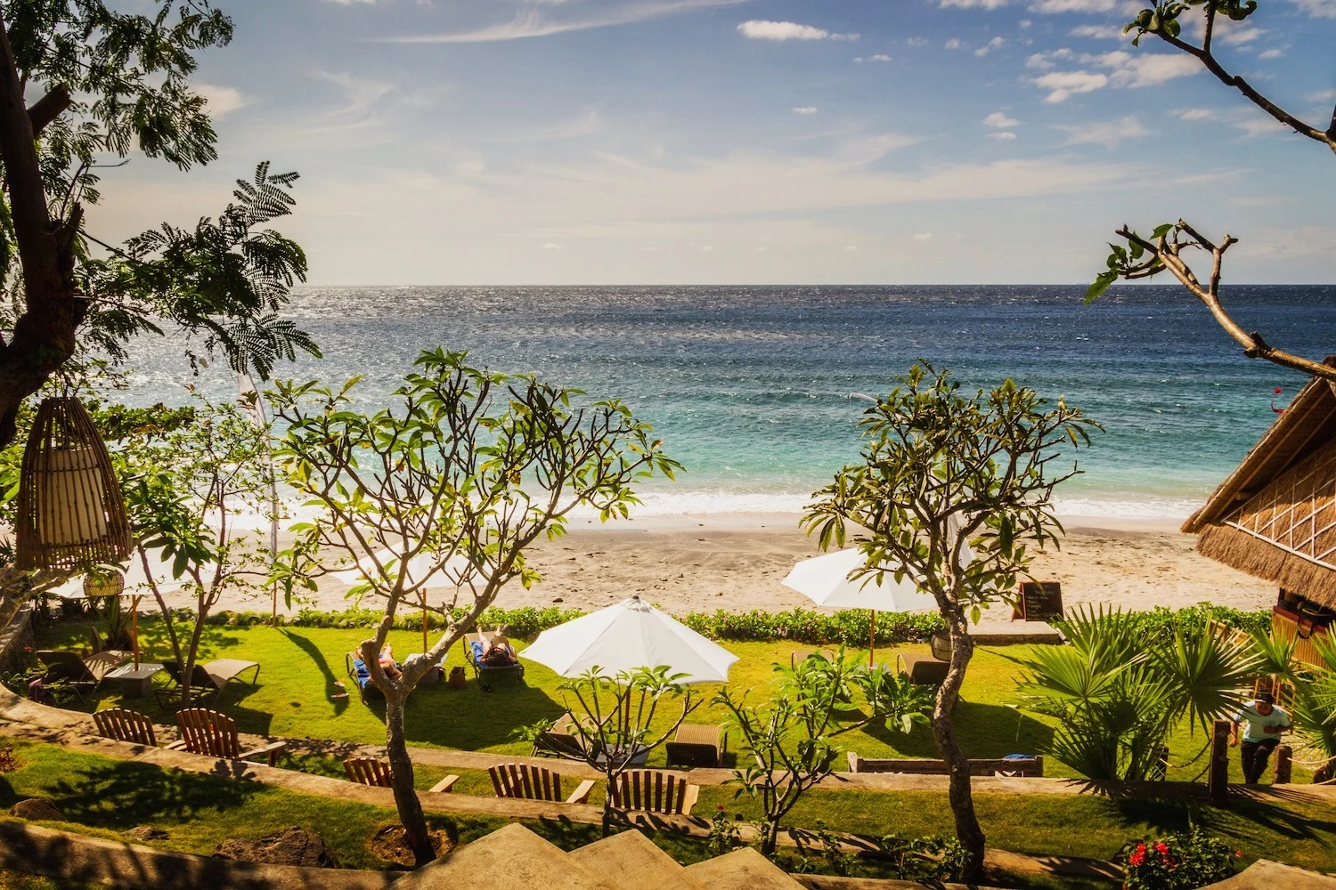 Lush green oceanfront garden with white sun loungers and umbrellas, tropical trees, and a sandy beach leading to turquoise water.